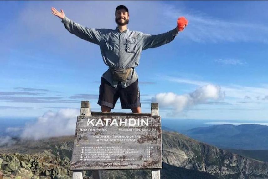 Hiker with arms outstretched on Katahdin summit sign, mountains in background.