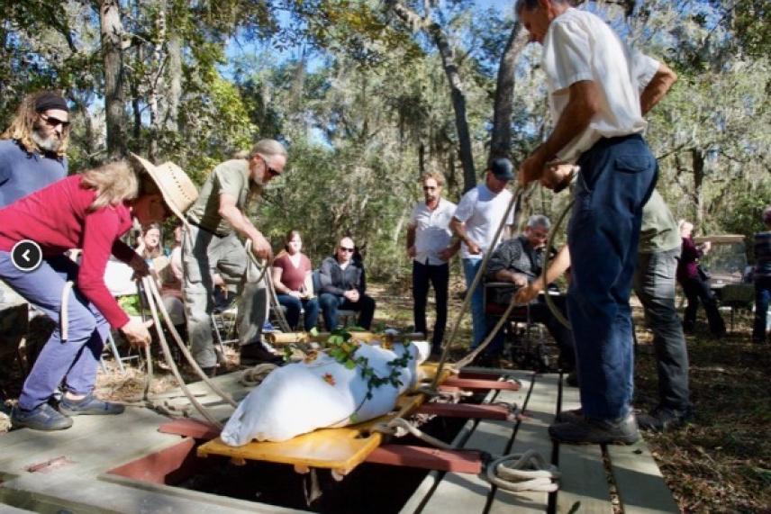 Group of people gather around a large alligator in a wooded area.