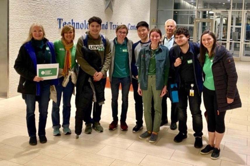 Group of smiling people standing indoors, some wearing green shirts.