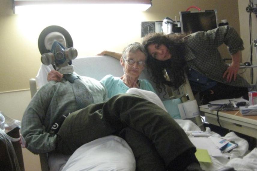 Hospital room with three people; one wearing a gas mask, others smiling.