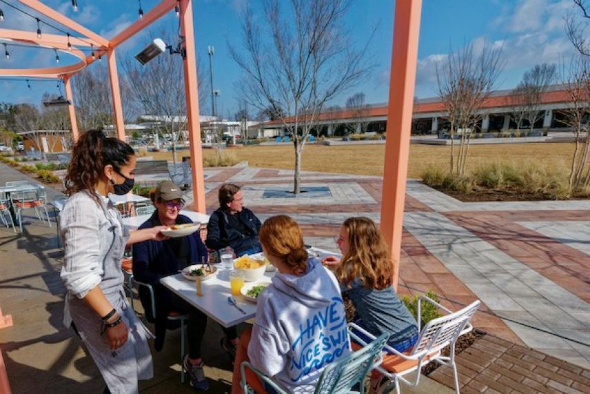 Server with a mask serves food to a group seated outdoors under a pergola.