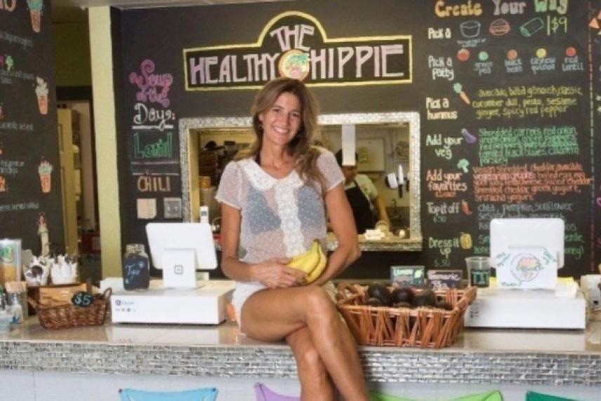 Woman seated on a counter smiling, holding bananas in a colorful juice bar setting.