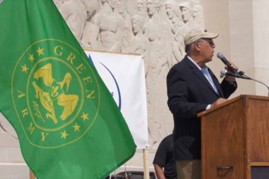 Man speaks at podium near green flag with emblem.