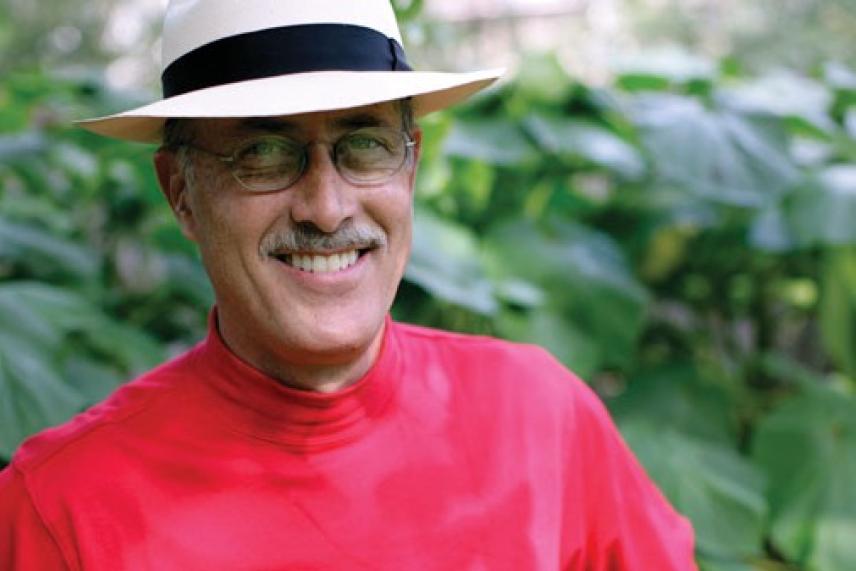 Smiling man in a white hat and red shirt, outdoors with green leaves in the background.