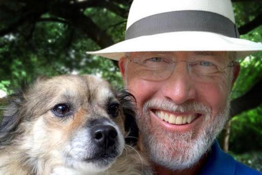 Smiling man in a hat with a small dog, greenery in the background.