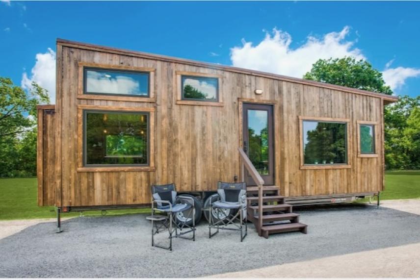 Tiny wooden house with large windows, a front porch, and outdoor seating on gravel.