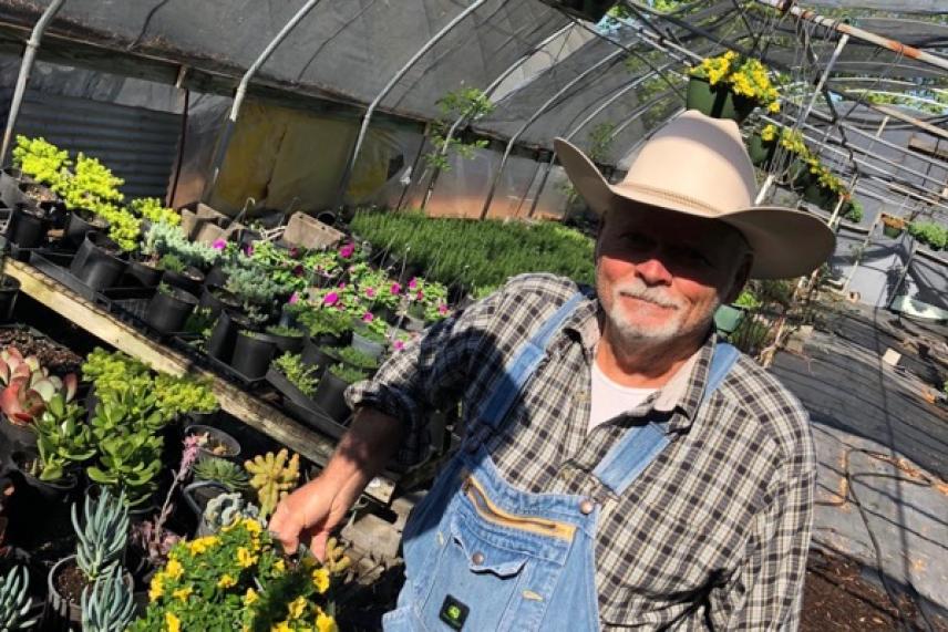 Man in a greenhouse wearing a hat and overalls, surrounded by plants.