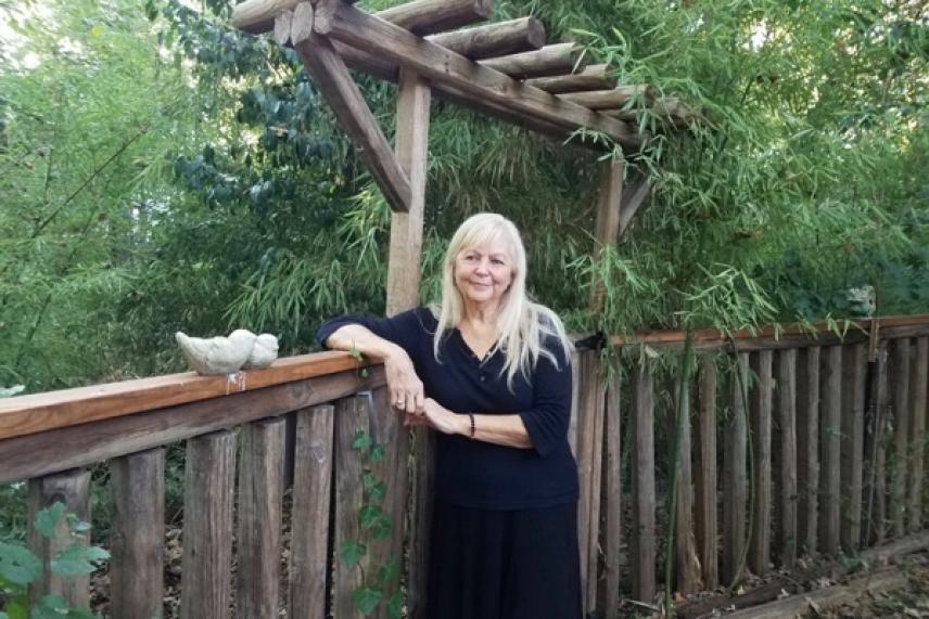Woman in black dress leaning on wooden fence in a garden setting.