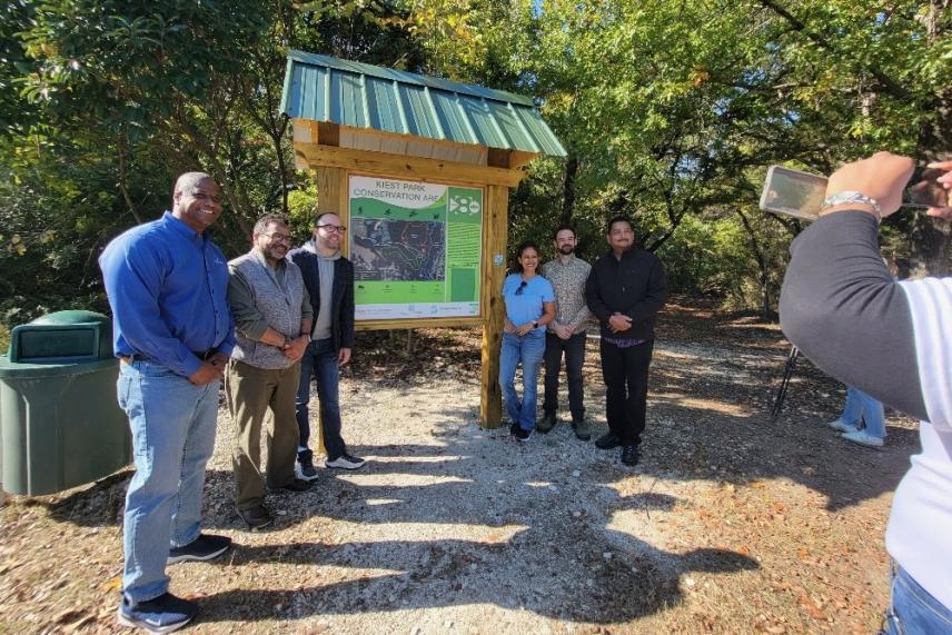 Group of people posing by a park information board, trees in the background.