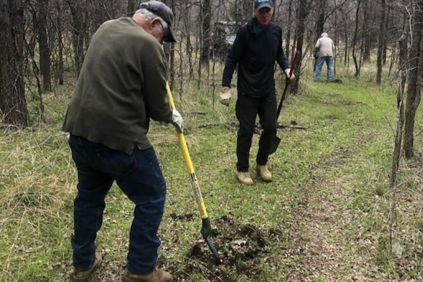 Two men digging in a wooded area, others in the background.