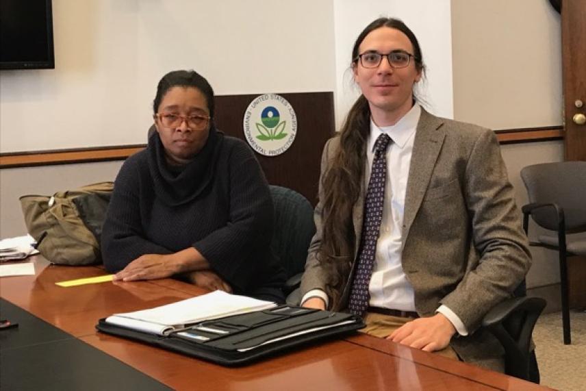 Two people seated at a conference table, one female, one male, with documents in front of them.