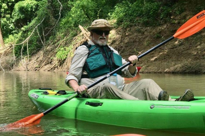 Man kayaking on a river in a green kayak, wearing a hat and sunglasses.