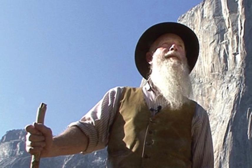 Man with long beard and hat holding a walking stick, standing outdoors against a rocky backdrop.