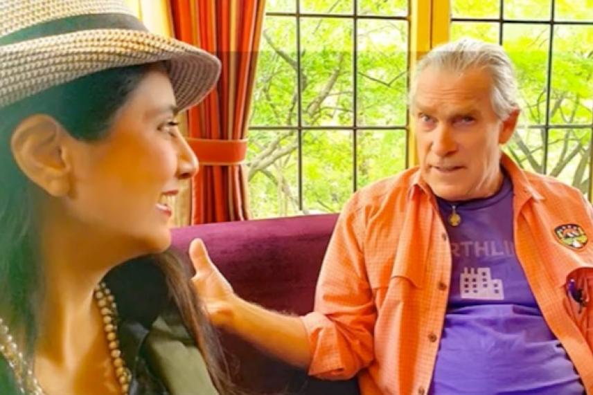 Two people talking on a couch near a window, one wearing a hat, the other in an orange shirt.