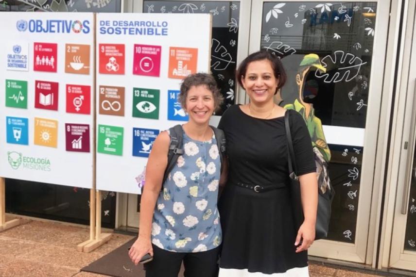 Two women smiling in front of sustainable development goals poster.