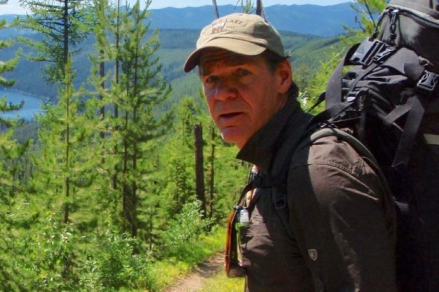 Man hiking on a forest trail, wearing a backpack and cap, with mountains in the background.