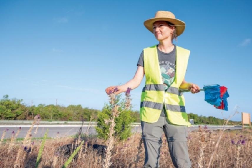 Worker in safety vest, straw hat holding flags, standing by a highway, clear sky.