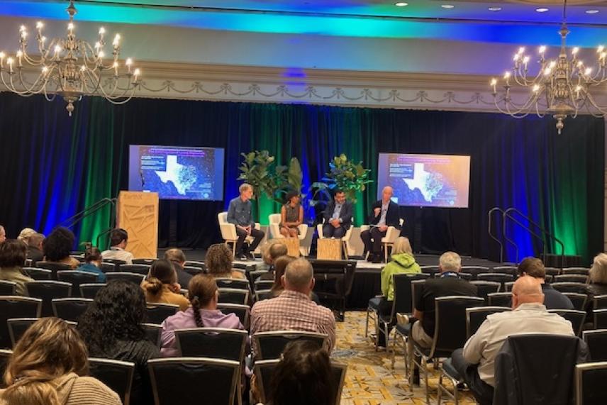 Panel of four speakers on stage at a conference, audience seated in the foreground.