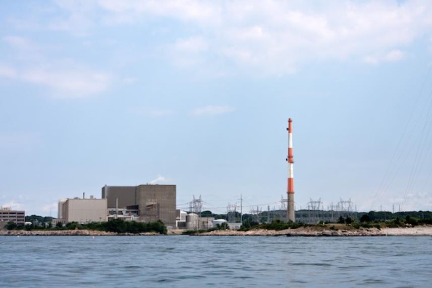 Industrial facility by water with a tall red and white chimney, under a cloudy sky.