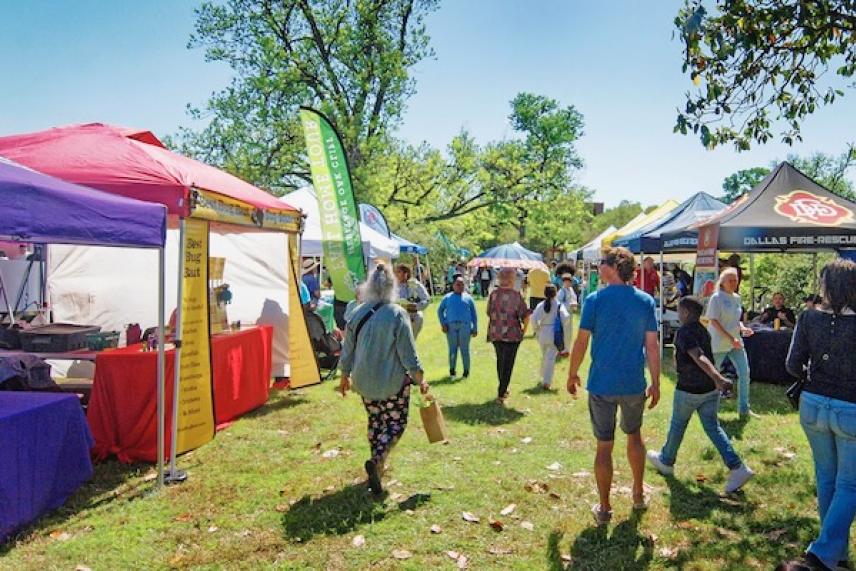 People walking between colorful market stalls on a sunny day in a park.