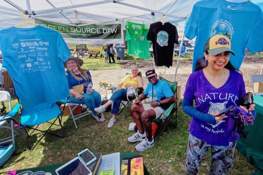 Festival booth with people sitting and standing, T-shirts hanging above.