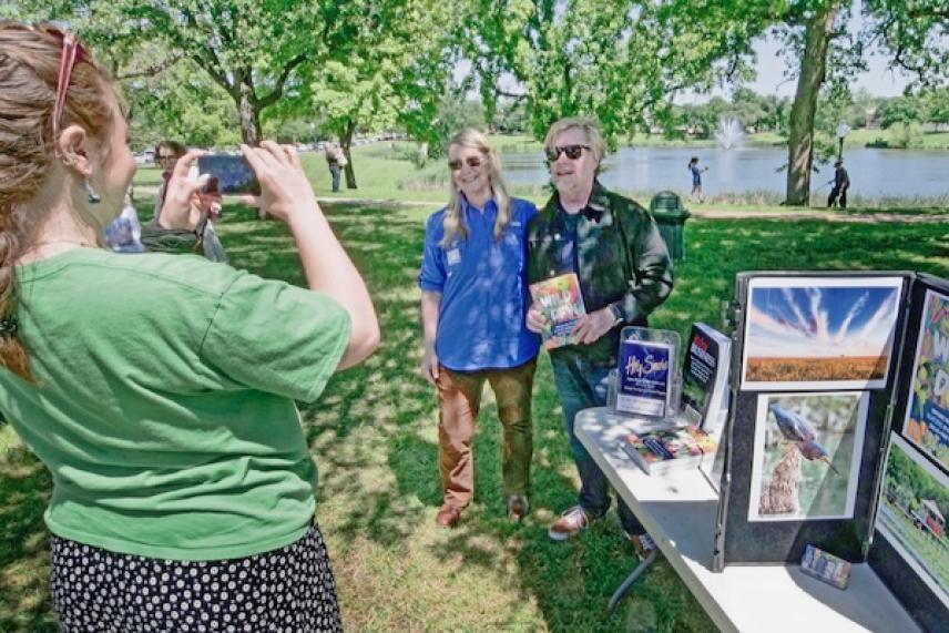 Person photographing two people at an outdoor event near a lake.