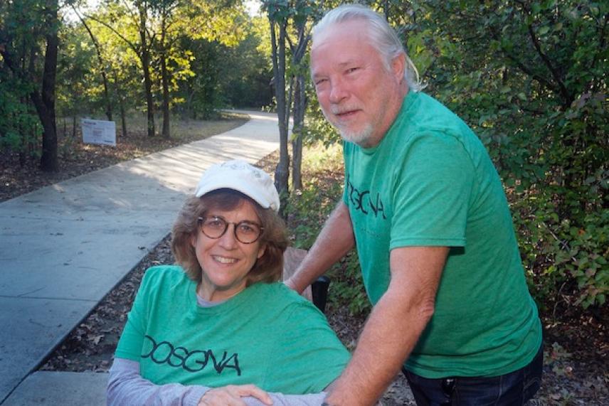 Smiling older couple in green shirts on a sunny forest pathway.