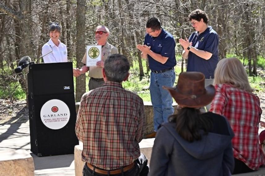 People applauding a speaker outside near trees, one holding an award.