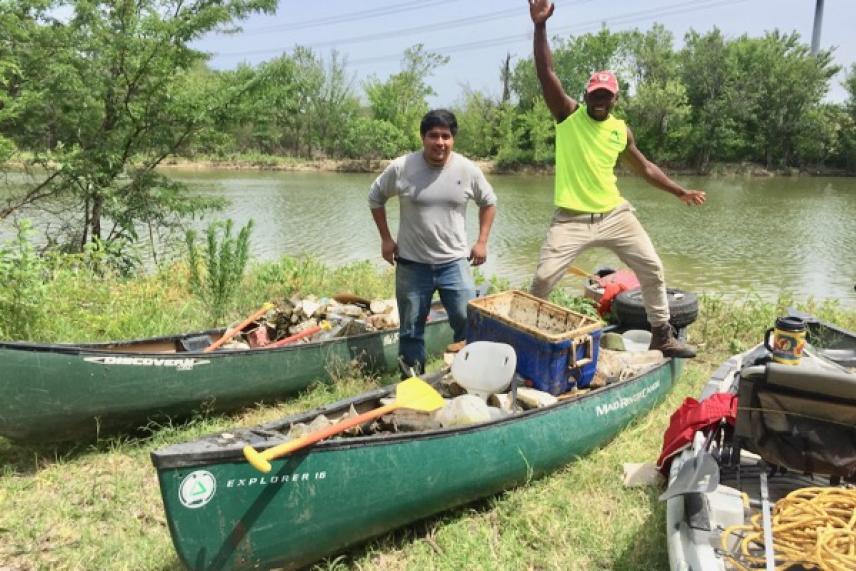 Two people with canoes filled with items by a riverbank on a sunny day.
