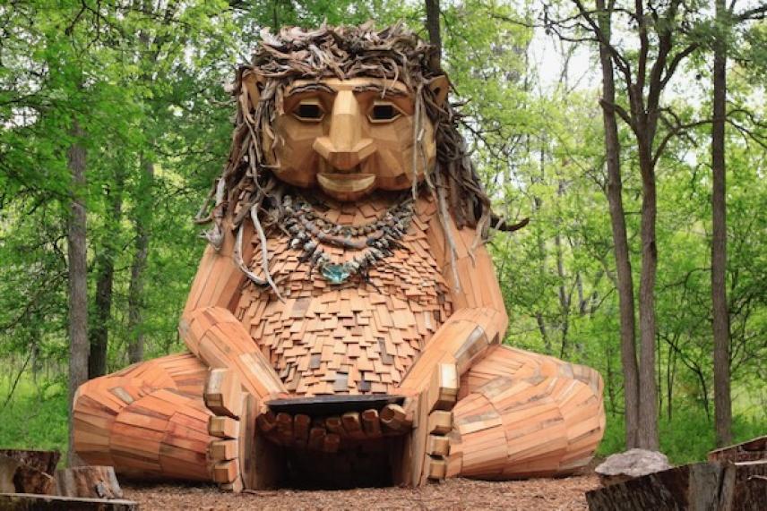 Wooden troll sculpture sitting in a forest clearing.