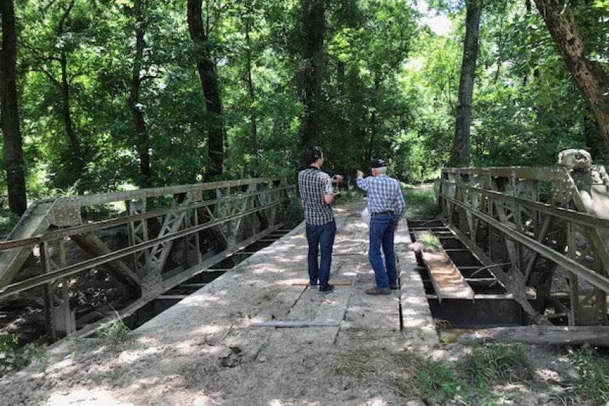 Two people stand on a narrow bridge in a forest setting.