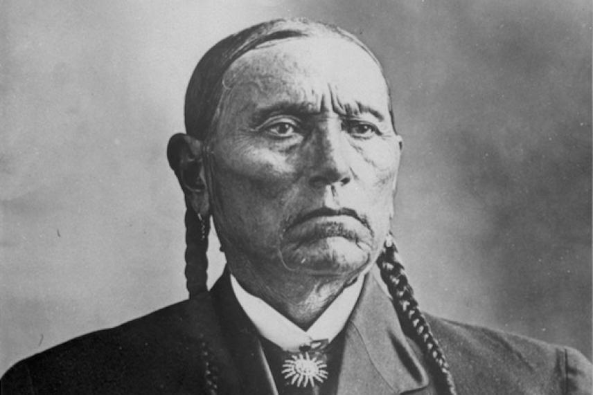 Elderly man with braided hair, wearing a suit and bolo tie, serious expression. Black and white photo.