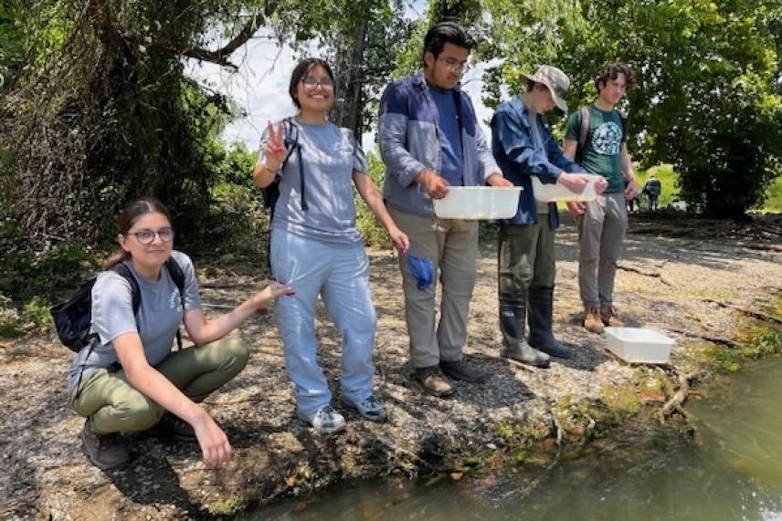 Five people engaged in water sampling by a riverbank.