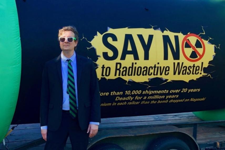 Man in suit and sunglasses stands beside anti-radioactive waste sign.