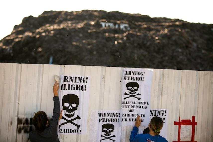 Two people place warning posters with skulls on a fence; mountain in the background.