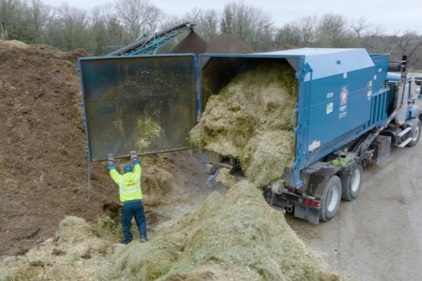 Truck offloading hay into a large pile, person in yellow vest nearby.