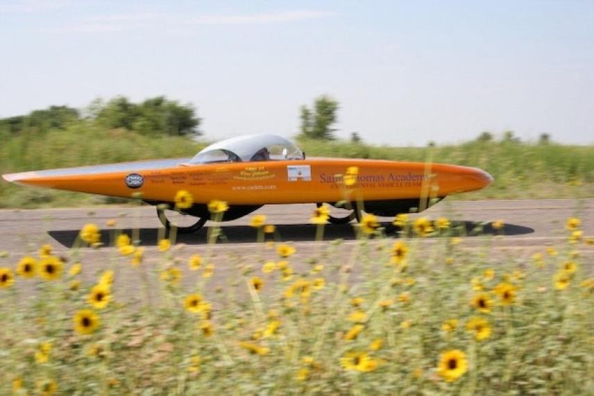 Orange solar car on a road, surrounded by yellow wildflowers.