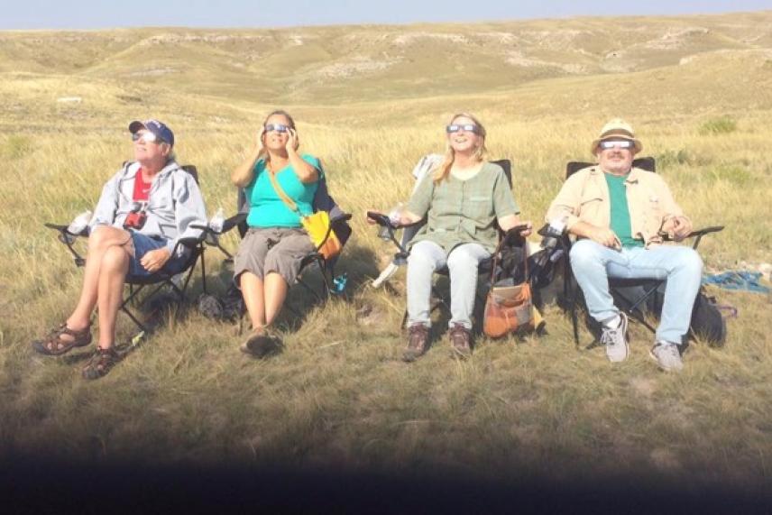 Four people in chairs on a grassy hill, wearing eclipse glasses.