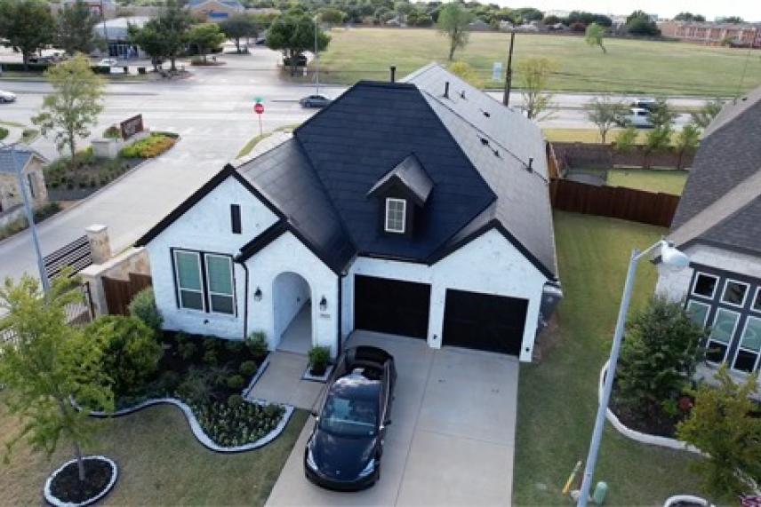 White house with black roof, two garages, and a car in the driveway, surrounded by greenery.