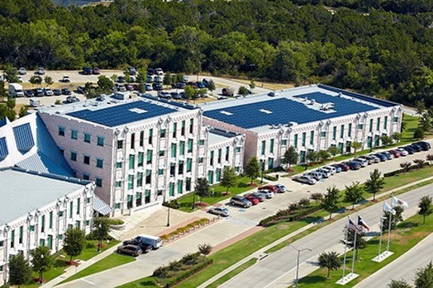 Large office building with solar panels and parked cars, surrounded by trees.