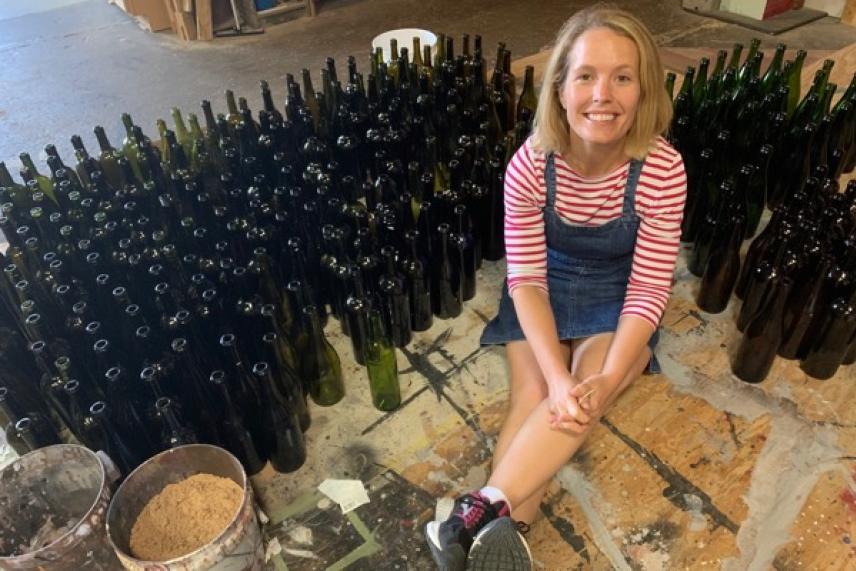 Smiling woman sitting on the floor, surrounded by numerous empty glass bottles.