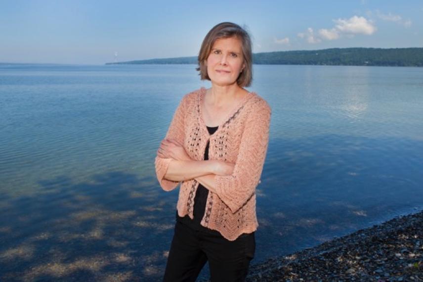 Woman standing by a calm lakeshore, arms crossed, under a clear sky.