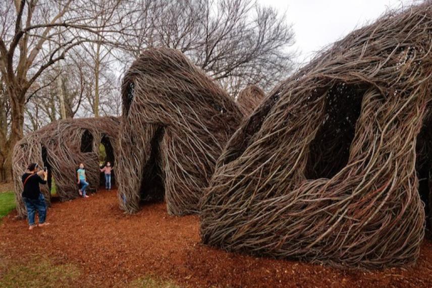 Twig sculptures shaped like huts on a grassy area with people nearby.