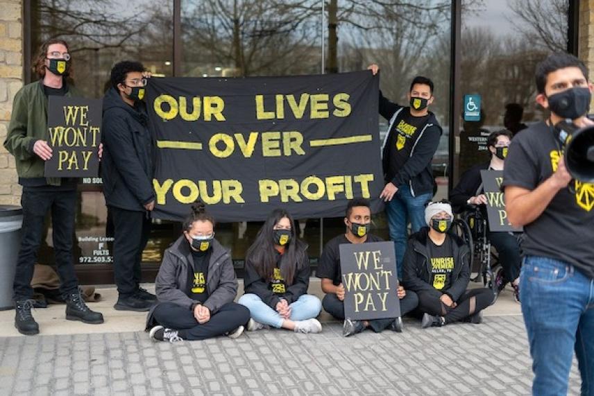 Protesters wearing masks hold signs about profit and lives, one speaks into a megaphone.