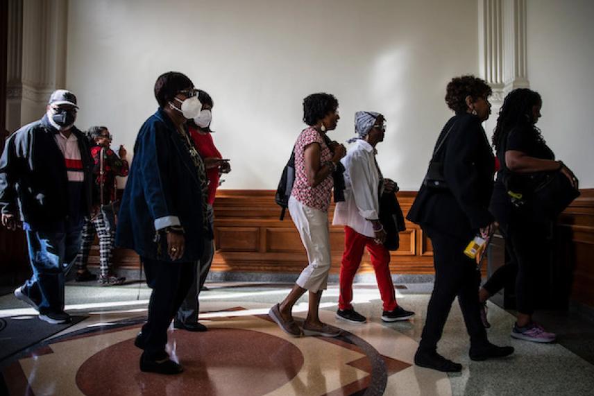 People walking in a sunlit hallway, some wearing masks.