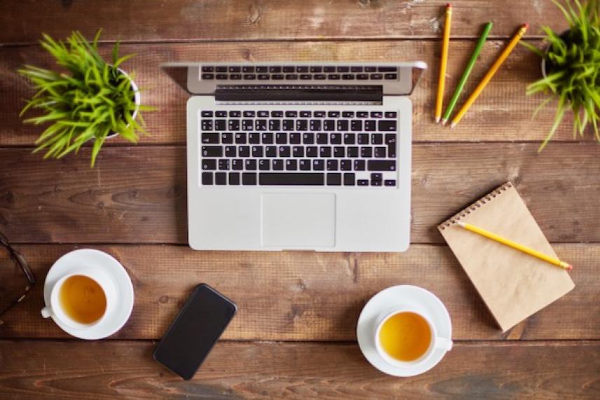 Laptop on wooden table with tea, phone, notebook, pencils, and small plants.