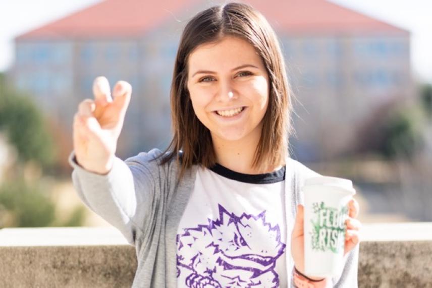 Smiling woman holding a coffee cup, gesturing with hand outdoors.