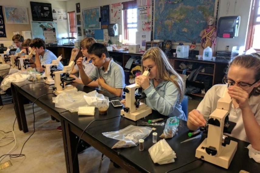Students in a classroom using microscopes on a long table.