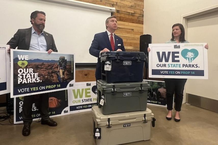 Three people hold signs promoting state parks inside a room with stacked coolers.