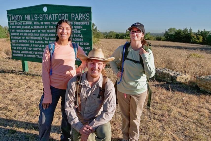 Three hikers smiling in a sunny, grassy park setting.